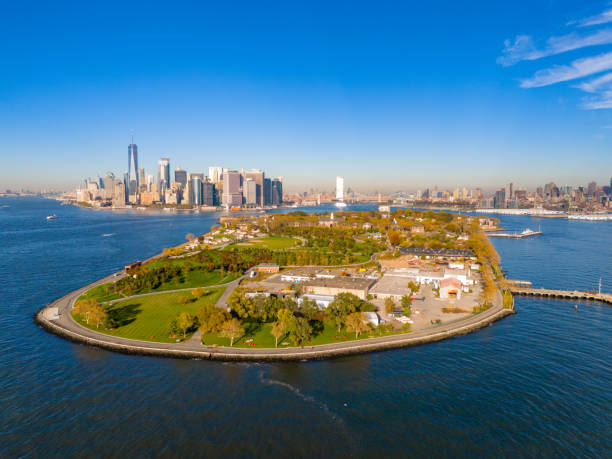 Aerial view of a grassy island with trees and buildings, surrounded by water, with the Manhattan skyline in the distance.