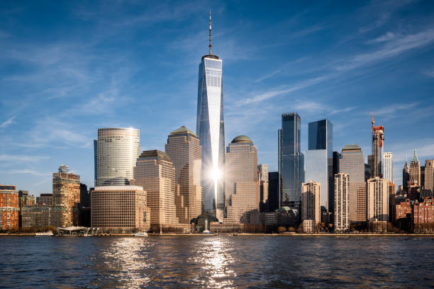 Skyline of Lower Manhattan with One World Trade Center, reflected on the river under a blue sky.