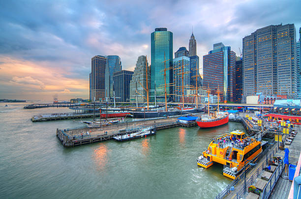 Manhattan waterfront with piers, a yellow ferry, and colorful boats against a skyline of tall glass buildings at dusk.