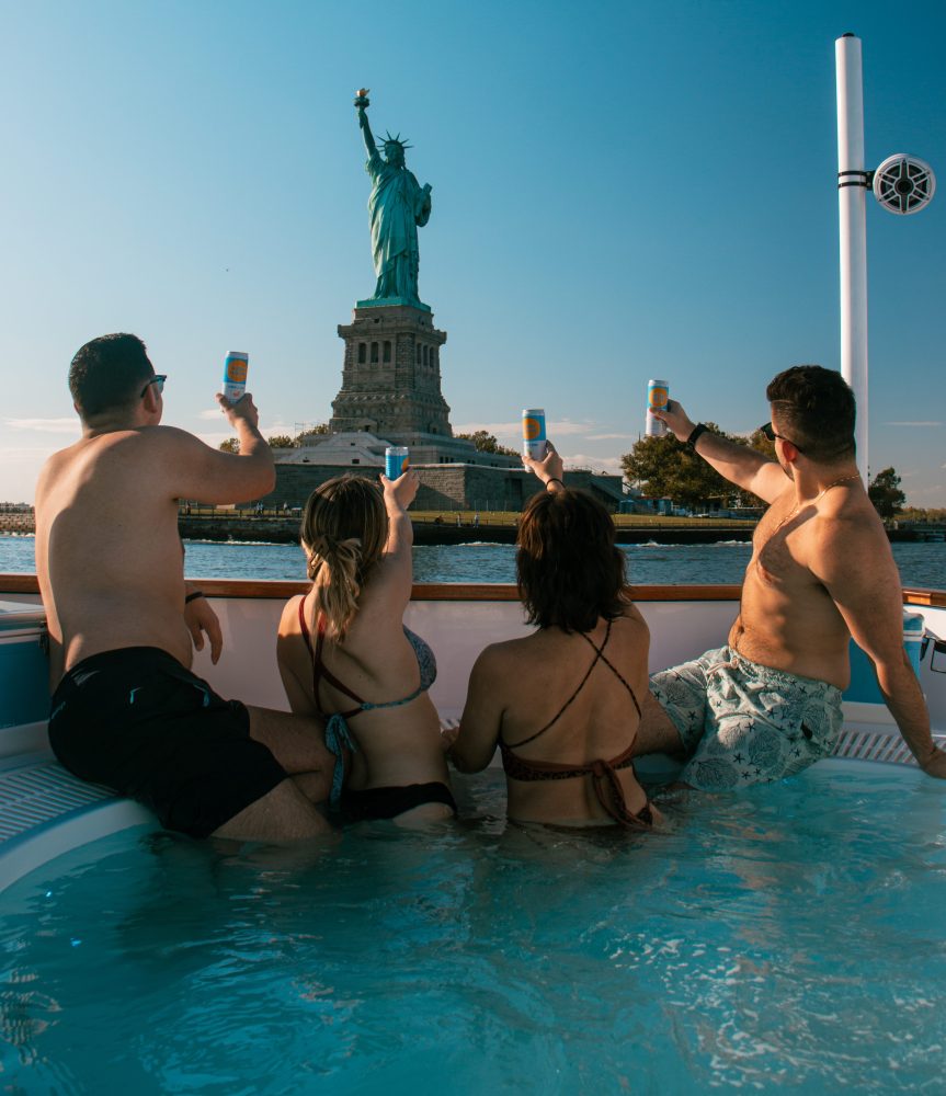 men and women in hot tub boat in front of statue of liberty