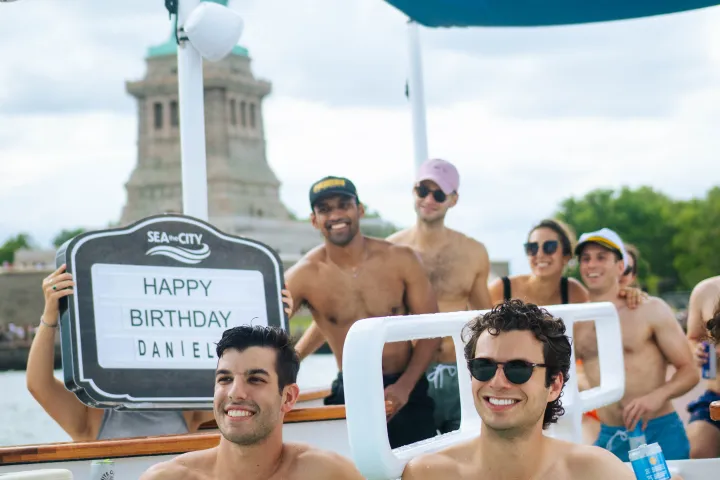 Group of friends in a boat hot tub, Statue of Liberty in background, holding birthday sign.