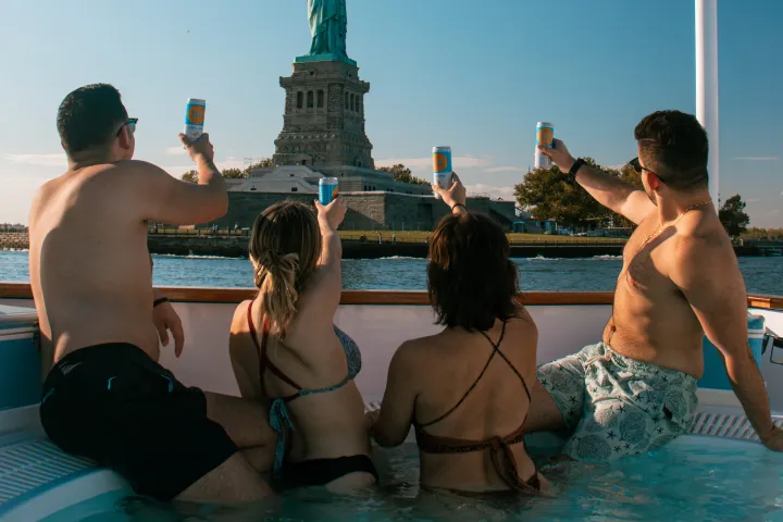 men and women in hot tub boat in front of statue of liberty