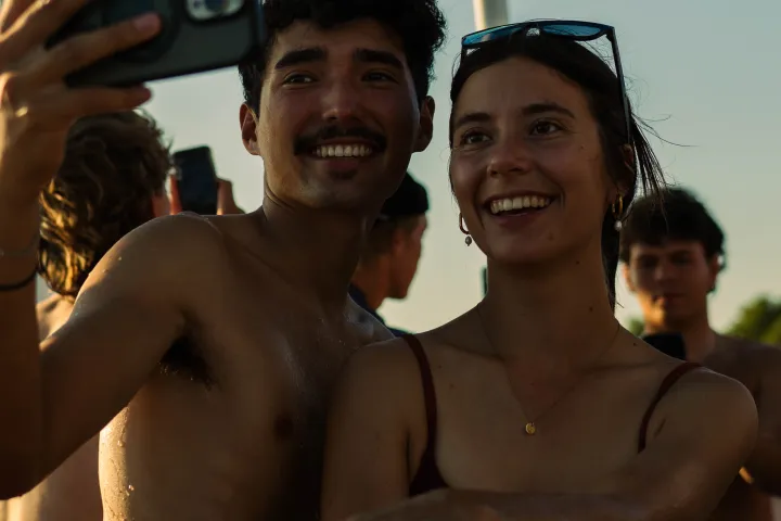 Smiling couple taking a selfie on a sunny day with others in background.