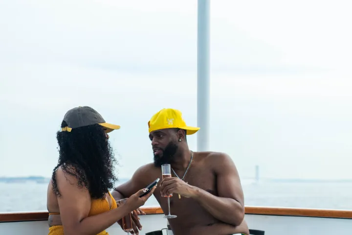 Two people sitting on a boat, chatting and holding drinks under a blue canopy.